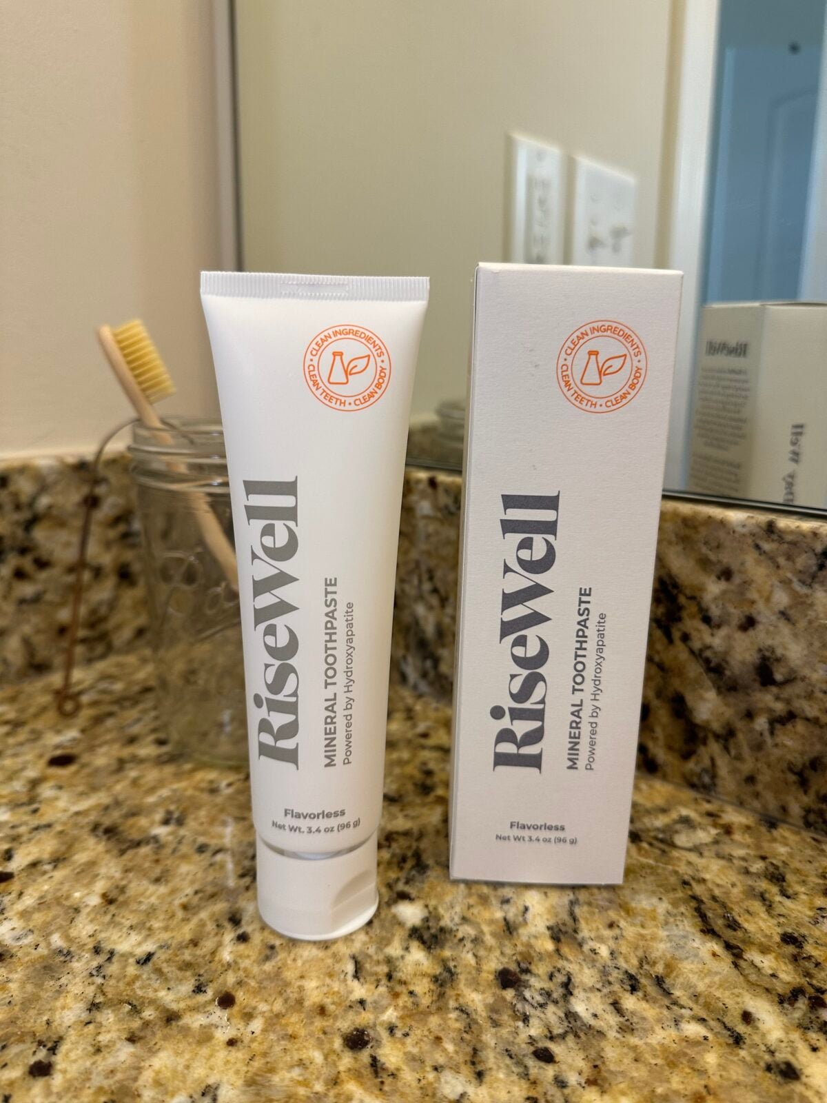 A tube and box of RiseWell mineral toothpaste, flavorless variety, sit on a granite bathroom countertop near a glass with a bamboo toothbrush. A mirror and electrical outlets are visible in the background.