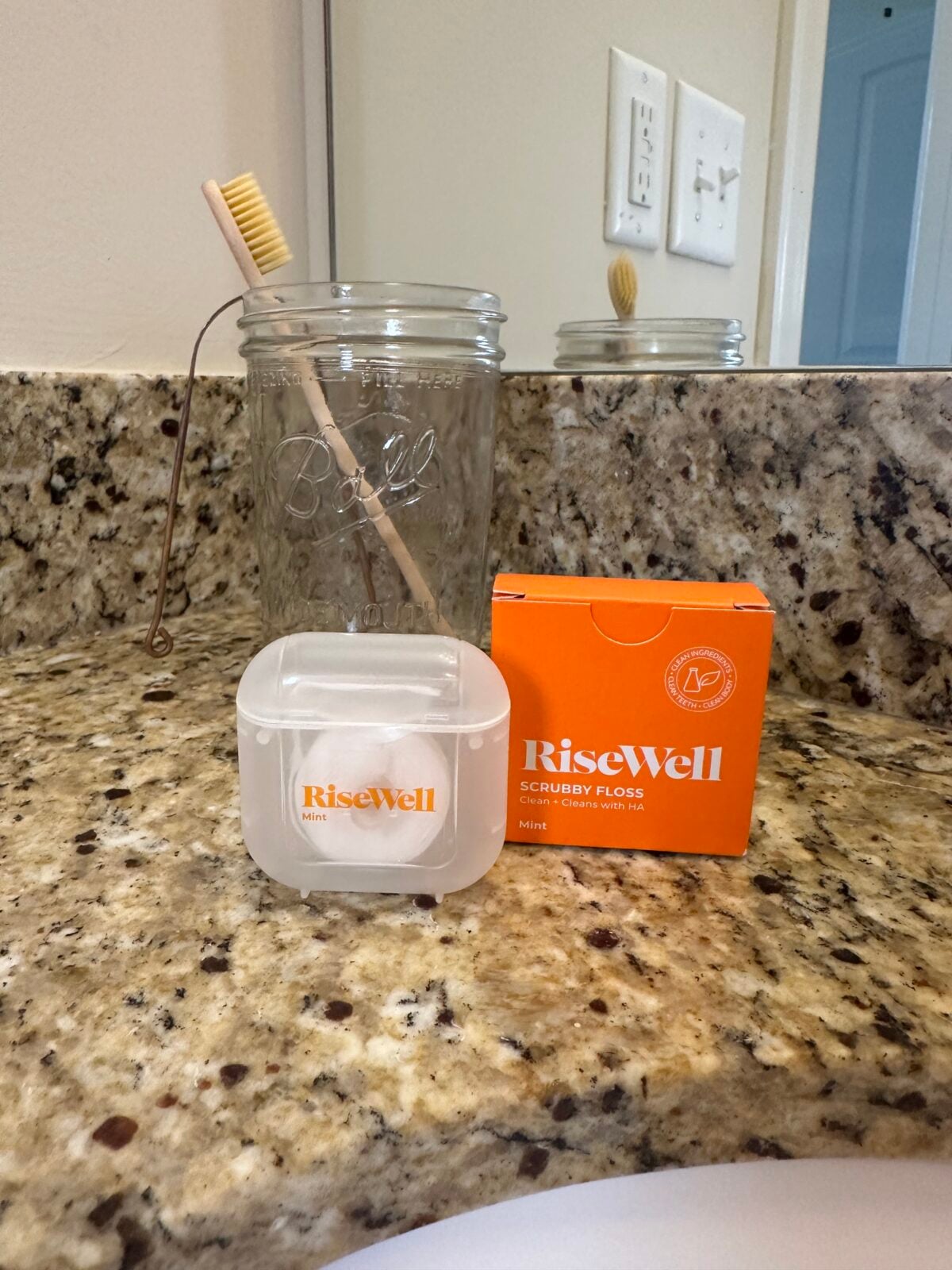 A bamboo toothbrush in a clear mason jar sits on a granite countertop next to a plastic container and orange box, both labeled &ldquo;RiseWell,&rdquo; in a bathroom setting.