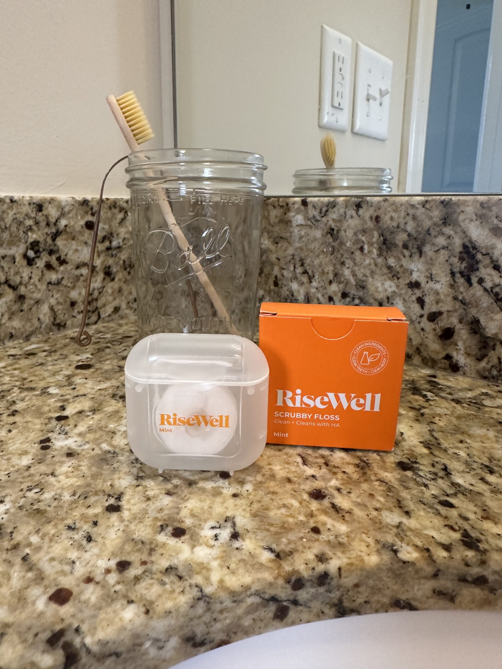 A bamboo toothbrush in a clear mason jar sits on a granite countertop next to a plastic container and orange box, both labeled &ldquo;RiseWell,&rdquo; in a bathroom setting.