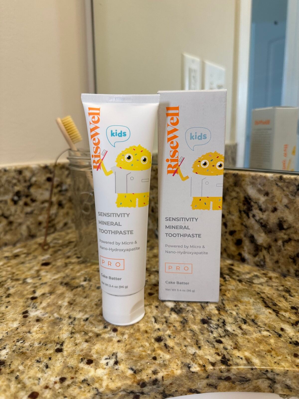 A tube and box of Risewell Kids Sensitivity Mineral Toothpaste sit on a granite bathroom counter. A brown toothbrush is in the background near a sink and a mirror.
