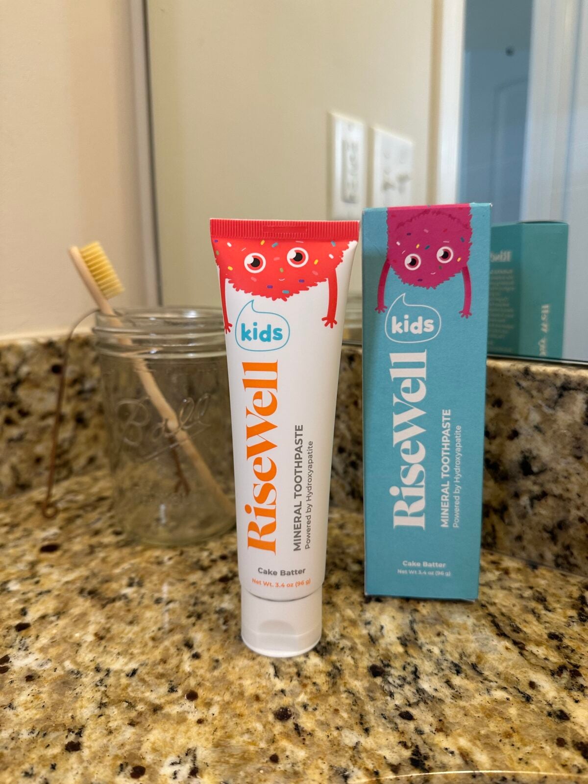 A tube of RiseWell Kids Mineral Toothpaste in Cake Batter flavor stands on a granite bathroom counter next to its box. A glass jar with a bamboo toothbrush is visible in the background.
