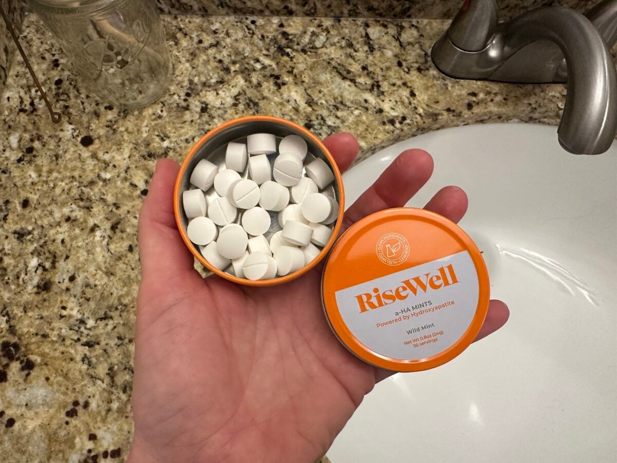 A hand holds an open orange container labeled RiseWell 3-in-1 Mints above a granite bathroom countertop next to a sink. The container is filled with round white mints.