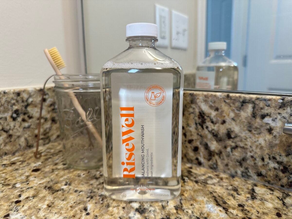 A clear plastic bottle of Risewell Balancing Mouthwash sits on a granite bathroom countertop next to a glass jar holding a wooden toothbrush. A mirror and light switches are visible in the background.