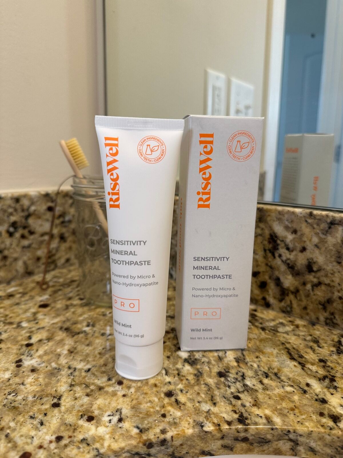 A tube and box of RiseWell Sensitivity Mineral Toothpaste are placed on a bathroom counter next to a glass jar holding a bamboo toothbrush. A mirror and a wall outlet are visible in the background.