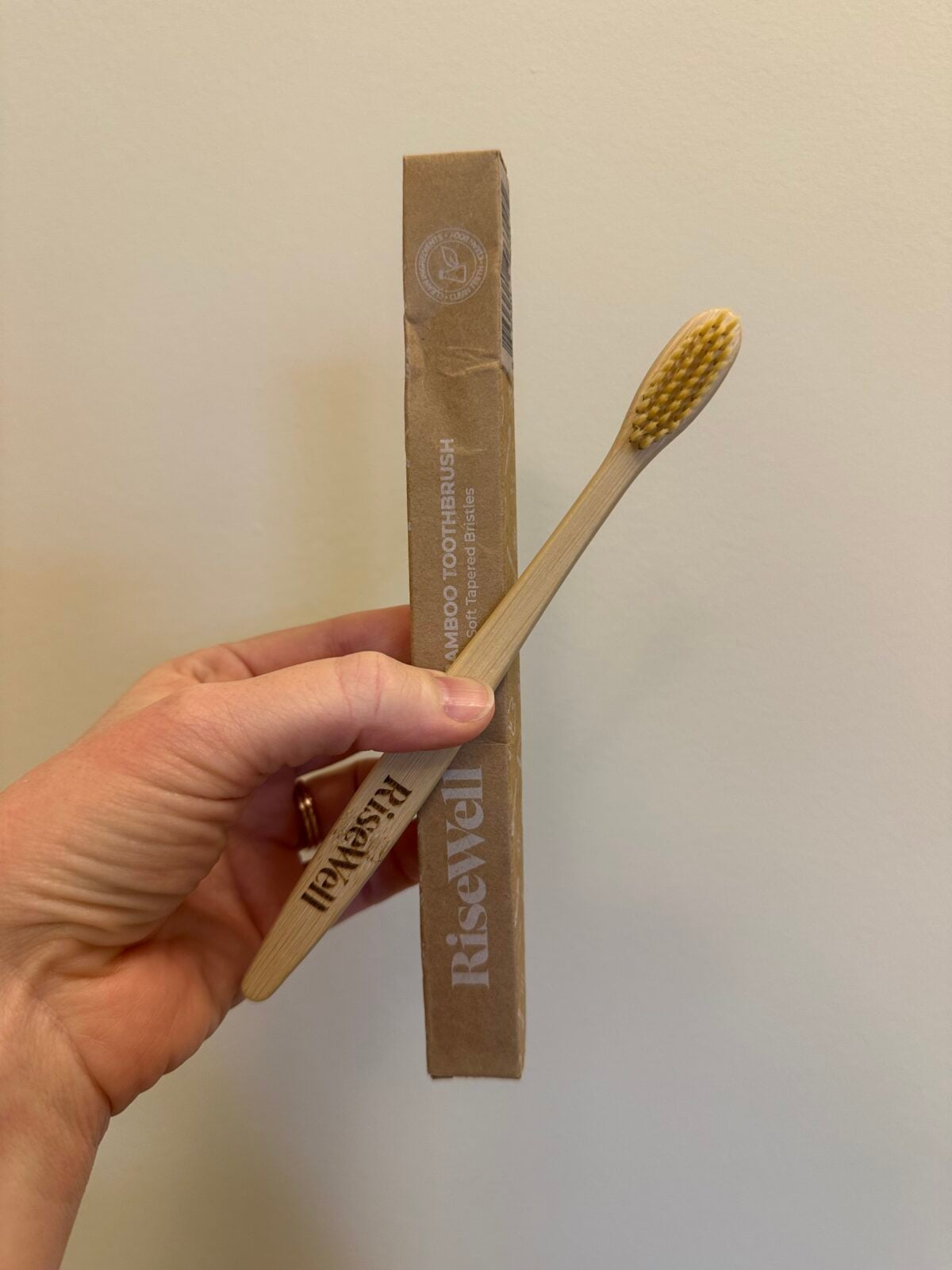 A hand holding a RiseWell bamboo toothbrush and its cardboard packaging against a plain, light-colored wall.