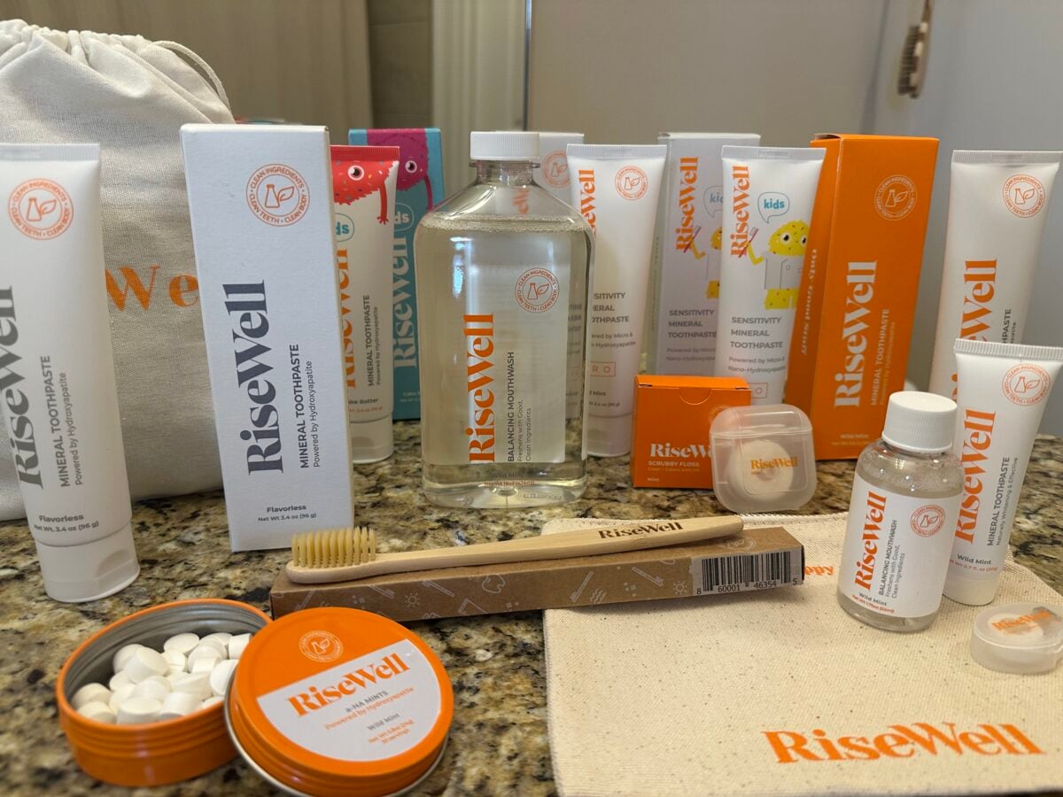 Various RiseWell oral care products, including toothpaste tubes, a mouthwash bottle, dental floss, a toothbrush, a container of tablets, and a drawstring bag, are arranged on a bathroom countertop.