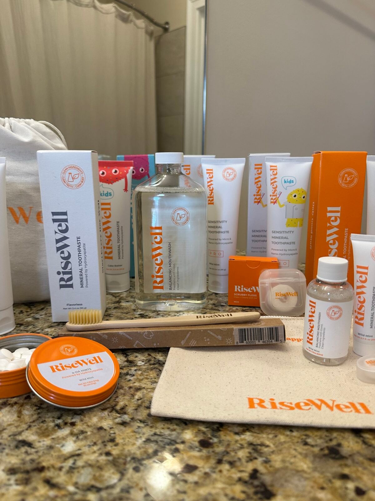 A variety of RiseWell oral care products, including toothpaste, mouthwash, floss, a toothbrush, chewable tablets, and a pouch, are arranged on a bathroom counter in front of a mirror.