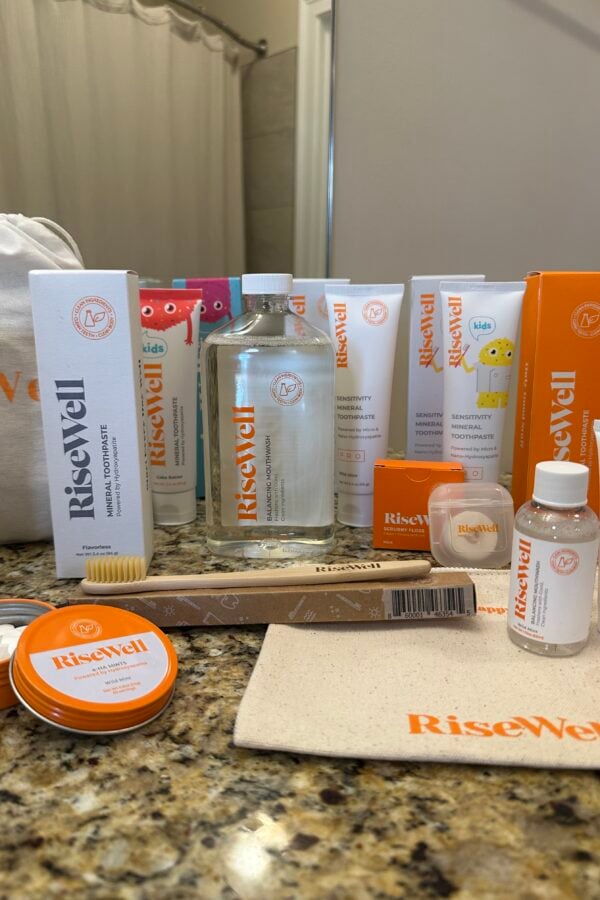 A variety of RiseWell oral care products, including toothpaste, mouthwash, floss, a toothbrush, chewable tablets, and a pouch, are arranged on a bathroom counter in front of a mirror.