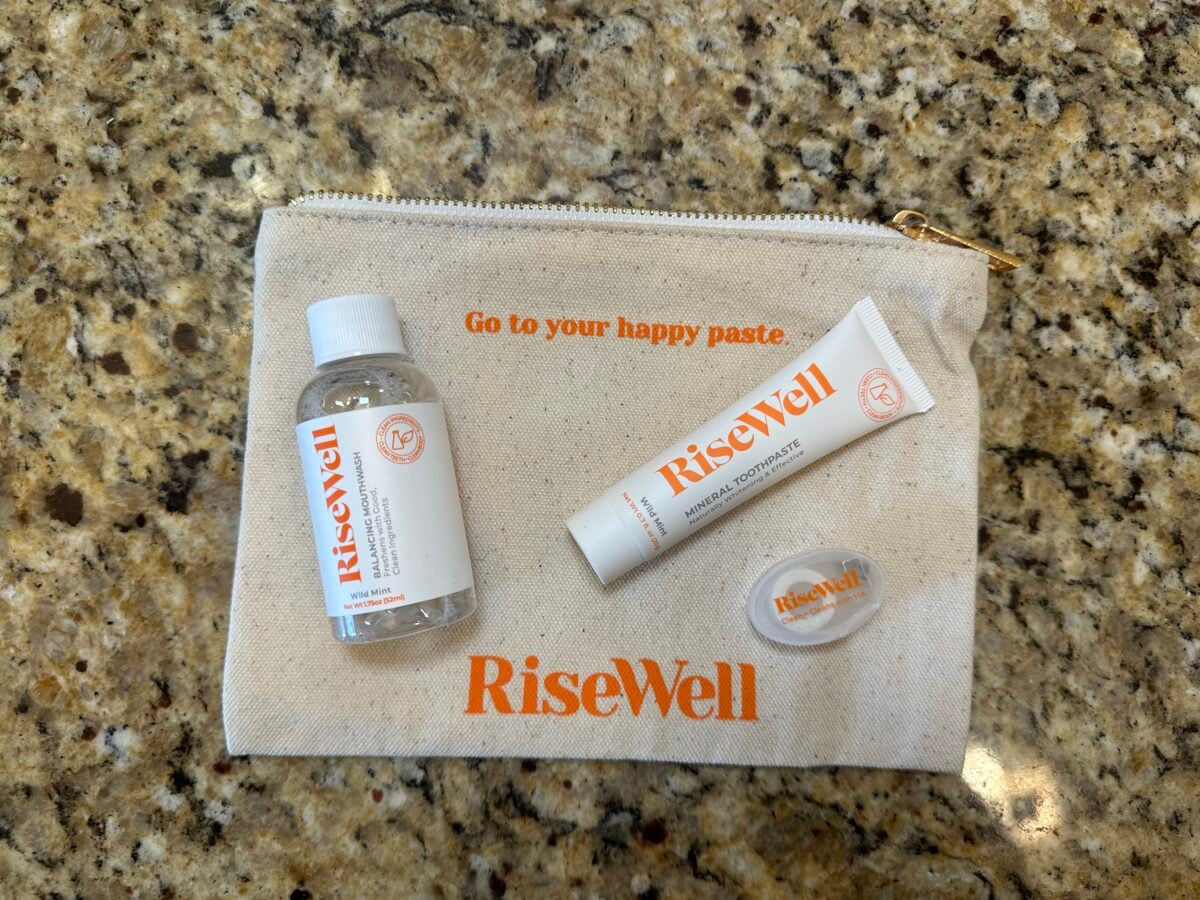 A small zippered pouch labeled &ldquo;RiseWell&rdquo; holds a travel-sized mouthwash bottle, a toothpaste tube, and a container of tooth chews, arranged on a granite countertop. The pouch reads, &ldquo;Go to your happy paste.&rdquo;.