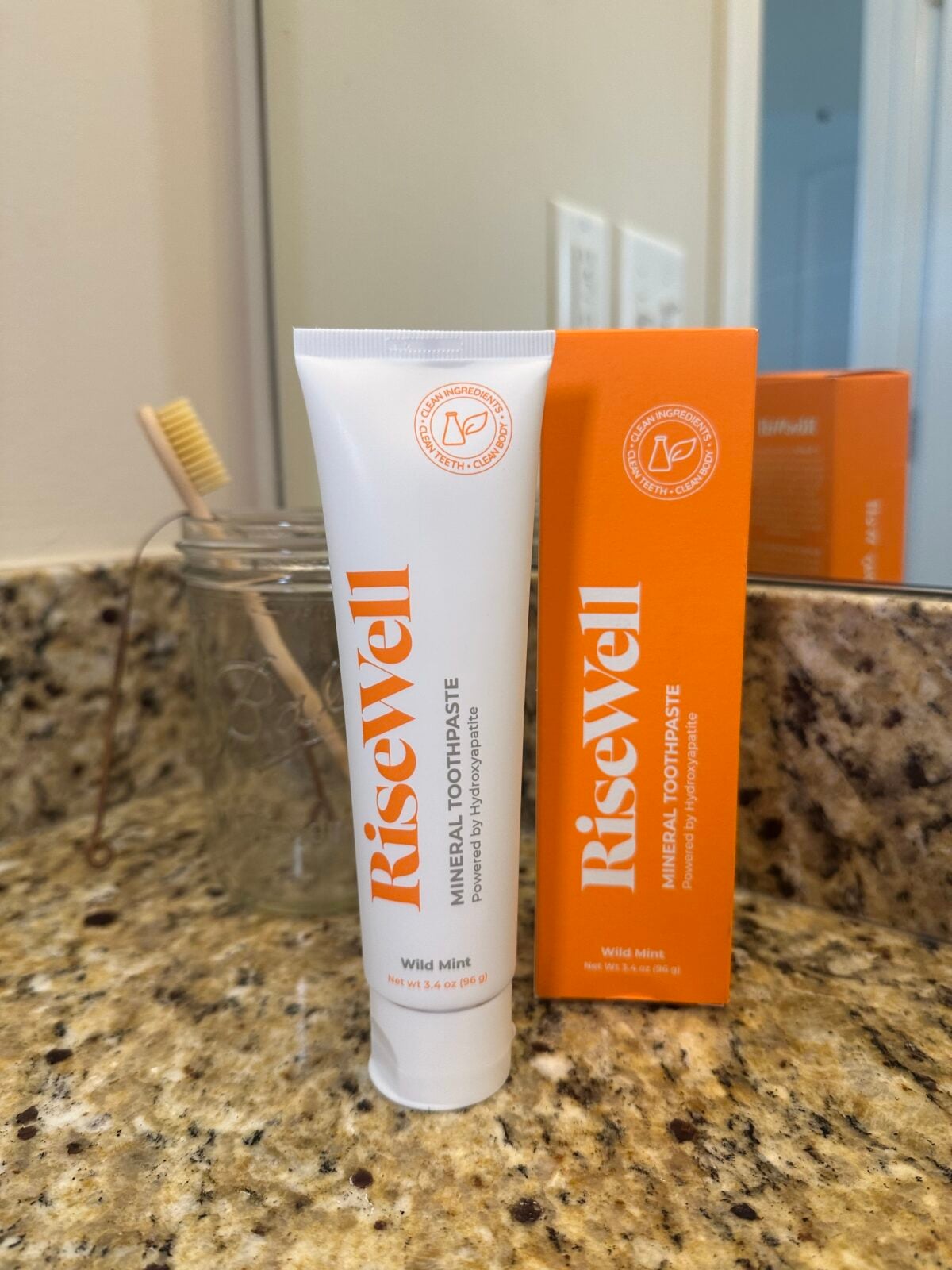 A tube of RiseWell Mineral Toothpaste in Wild Mint flavor stands upright next to its orange box on a bathroom counter. A glass jar with a bamboo toothbrush is visible in the background.