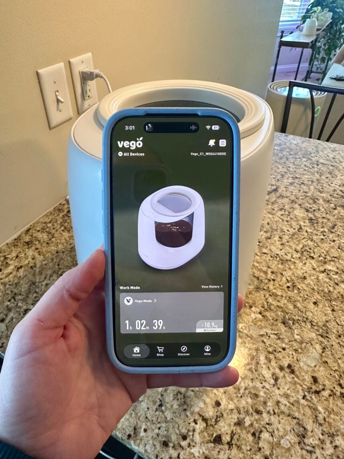 A hand holds a smartphone displaying the Vegō app with an image of a white kitchen appliance that matches the device on the granite countertop in the background. The app shows a timer and device status.
