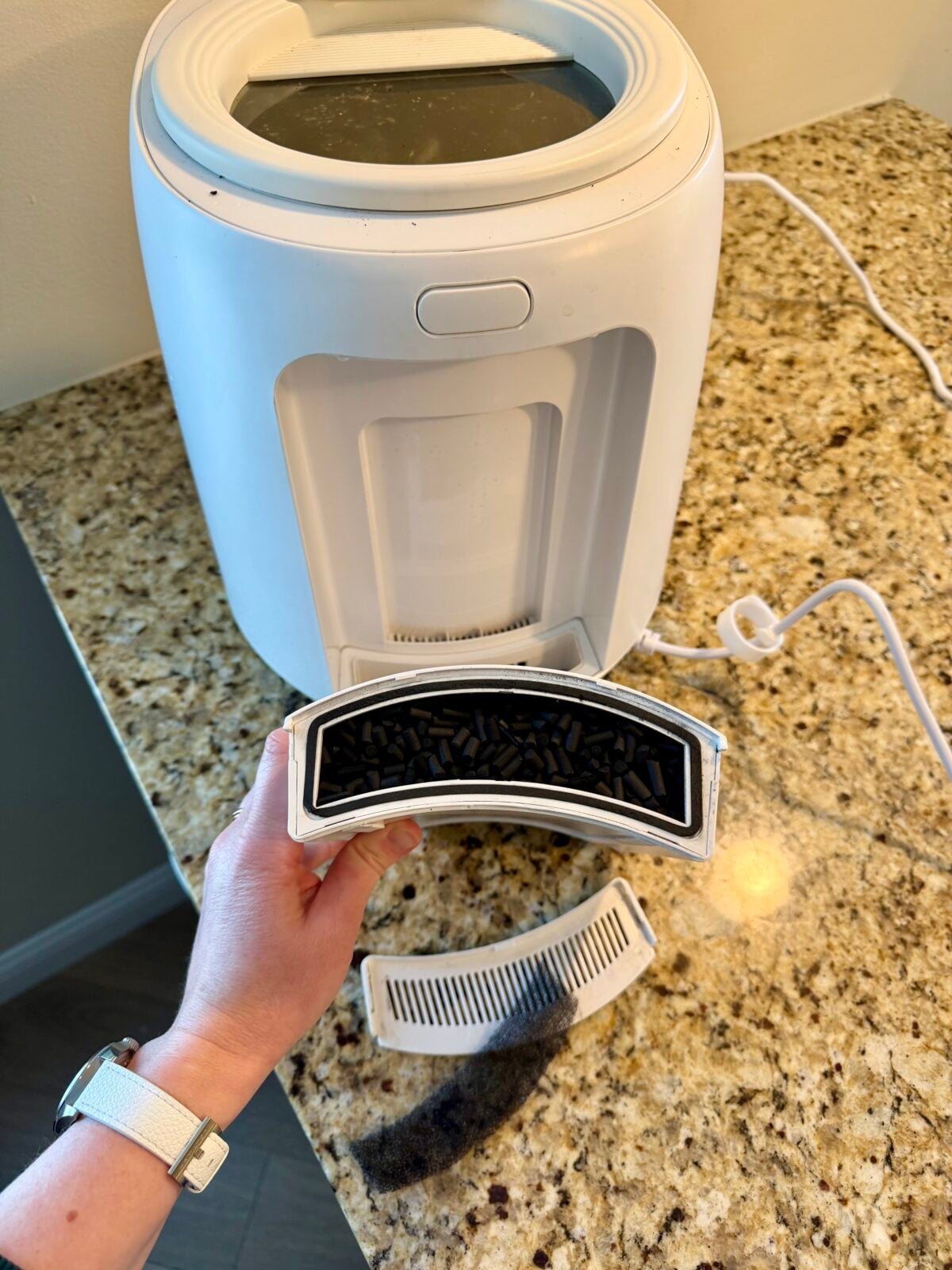 A hand is holding a removable filter with trapped pet hair and debris in front of a white automatic pet litter box on a granite countertop. The litter box is plugged in, and the filter appears recently cleaned.