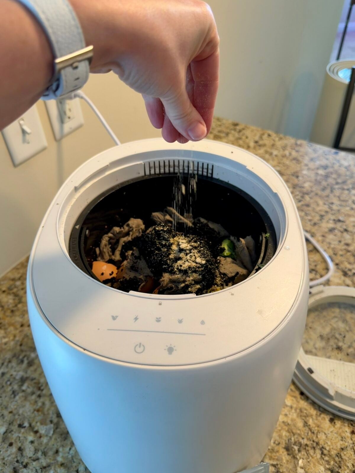 A hand sprinkles powder into a food composter filled with food scraps, sitting on a kitchen counter. The composter has a white exterior and touch controls on the front.