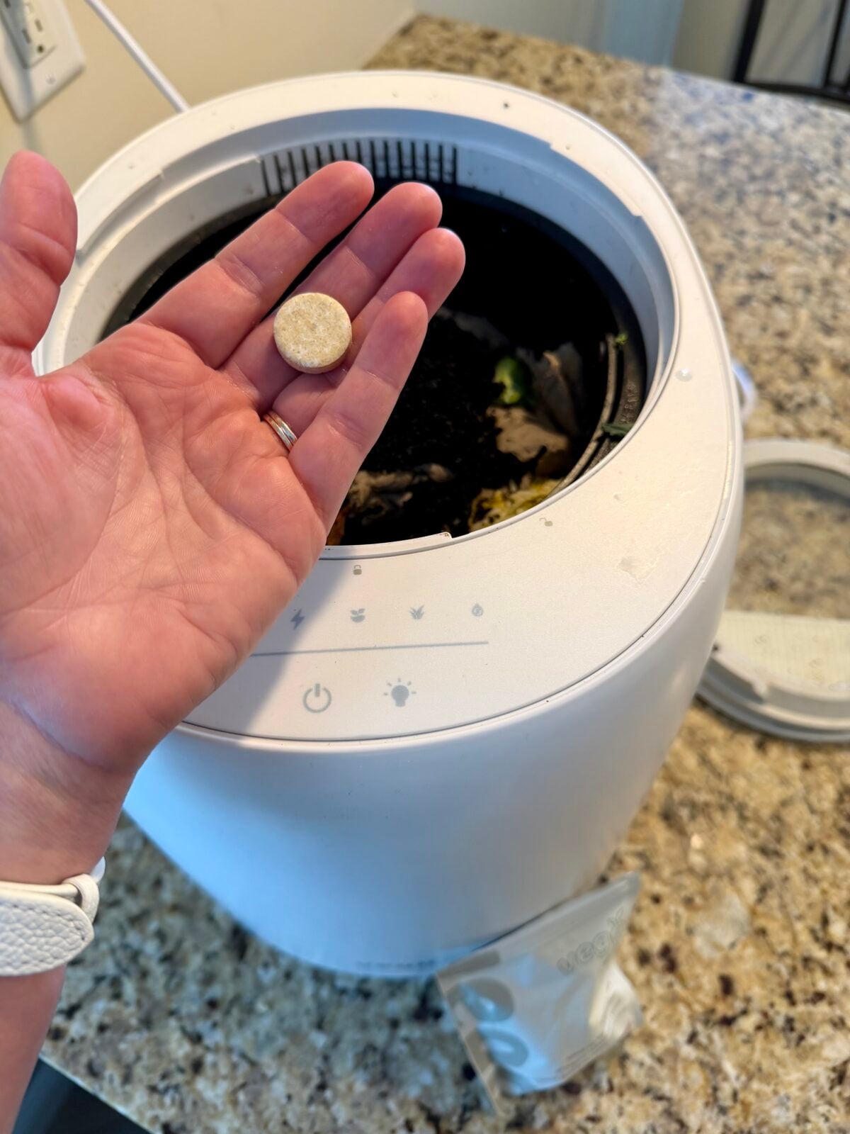 A person holds a small round tablet above an open white countertop compost bin, which contains food scraps. The compost bin has control buttons on the lid and sits on a speckled countertop.