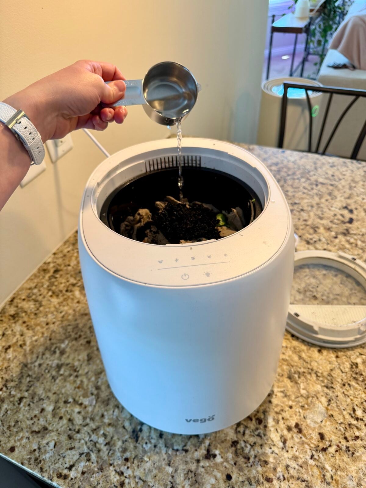 A hand pours water from a metal measuring cup into a white kitchen compost bin filled with food scraps, positioned on a granite countertop.