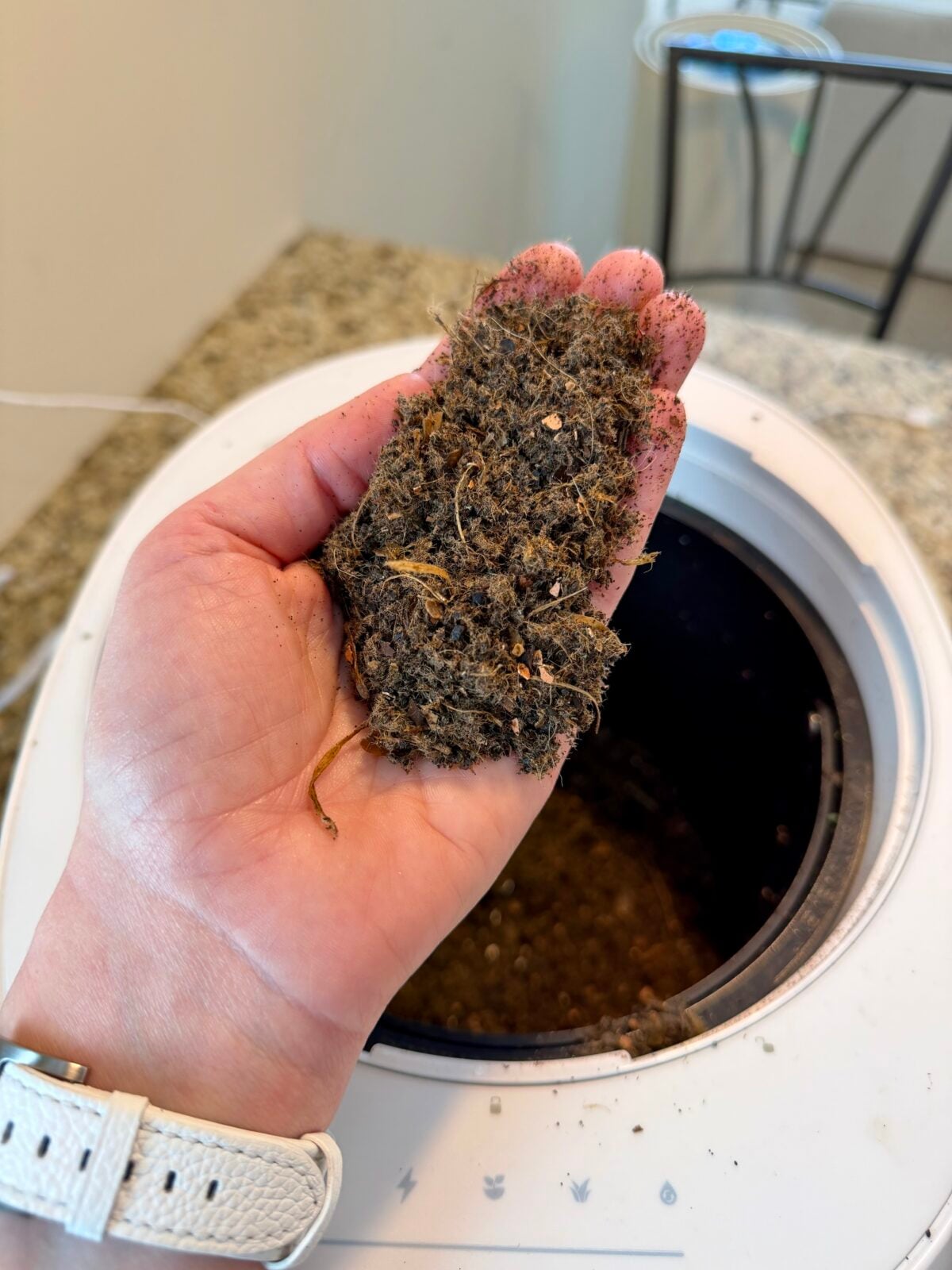 A hand holds a small pile of dark, crumbly compost above an open compost bin on a kitchen counter. The compost contains visible bits of organic matter.