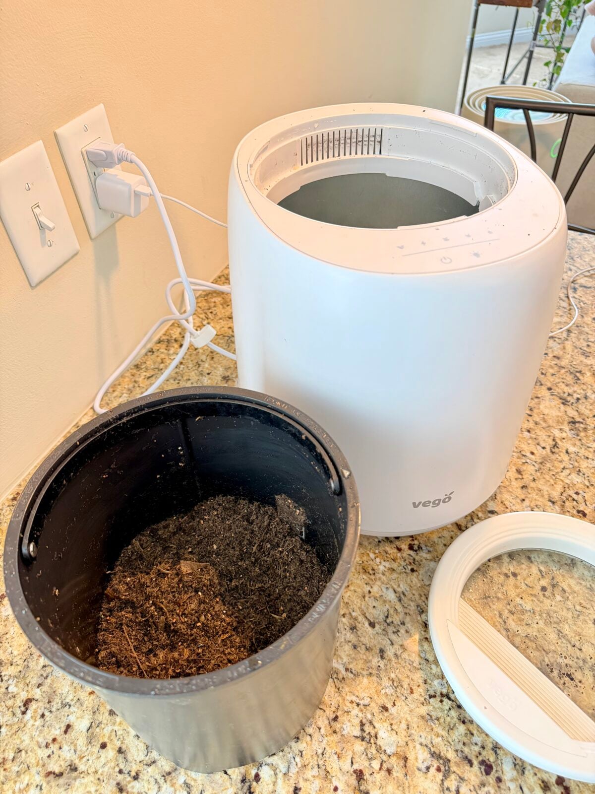 A white electric kitchen composter sits on a granite countertop, plugged into the wall. Its black inner bin is removed and filled with processed compost, and the lid rests nearby.