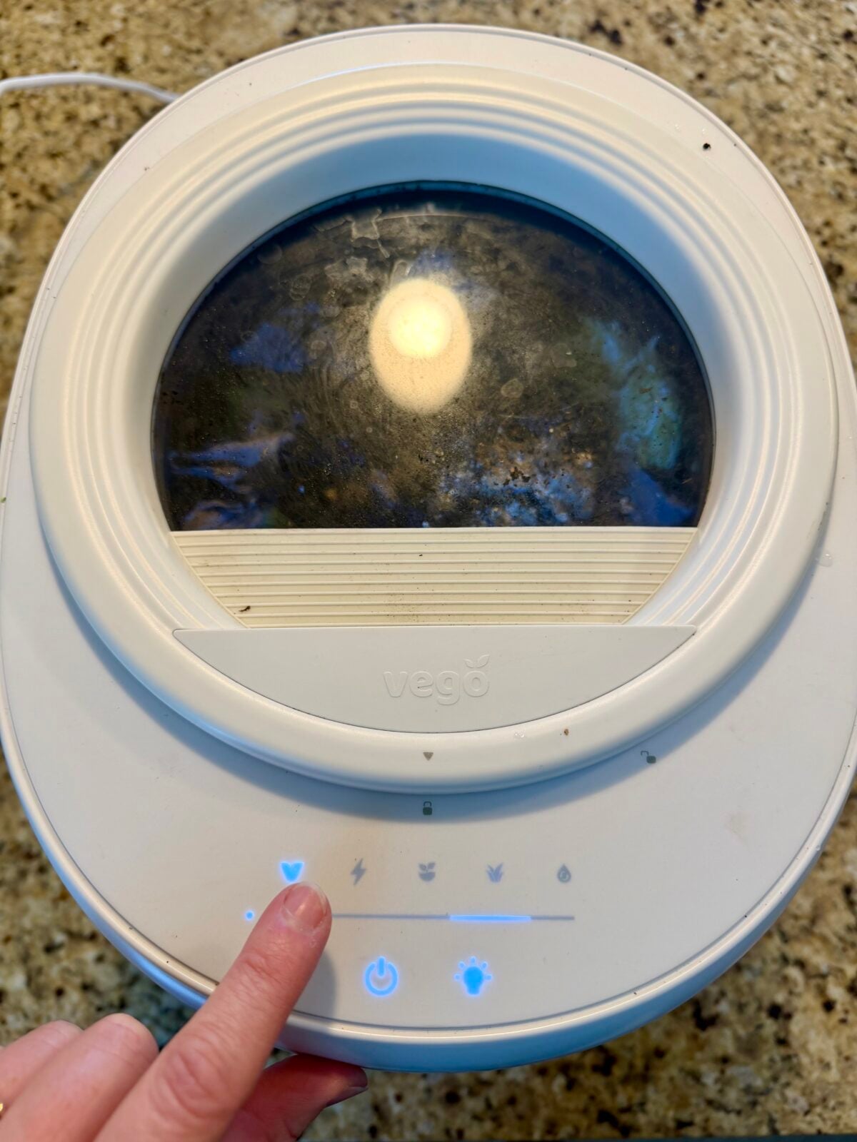 A hand presses a button on a white countertop appliance labeled vego with a round windowed lid. The control panel has illuminated icons, and the appliance sits on a speckled countertop.