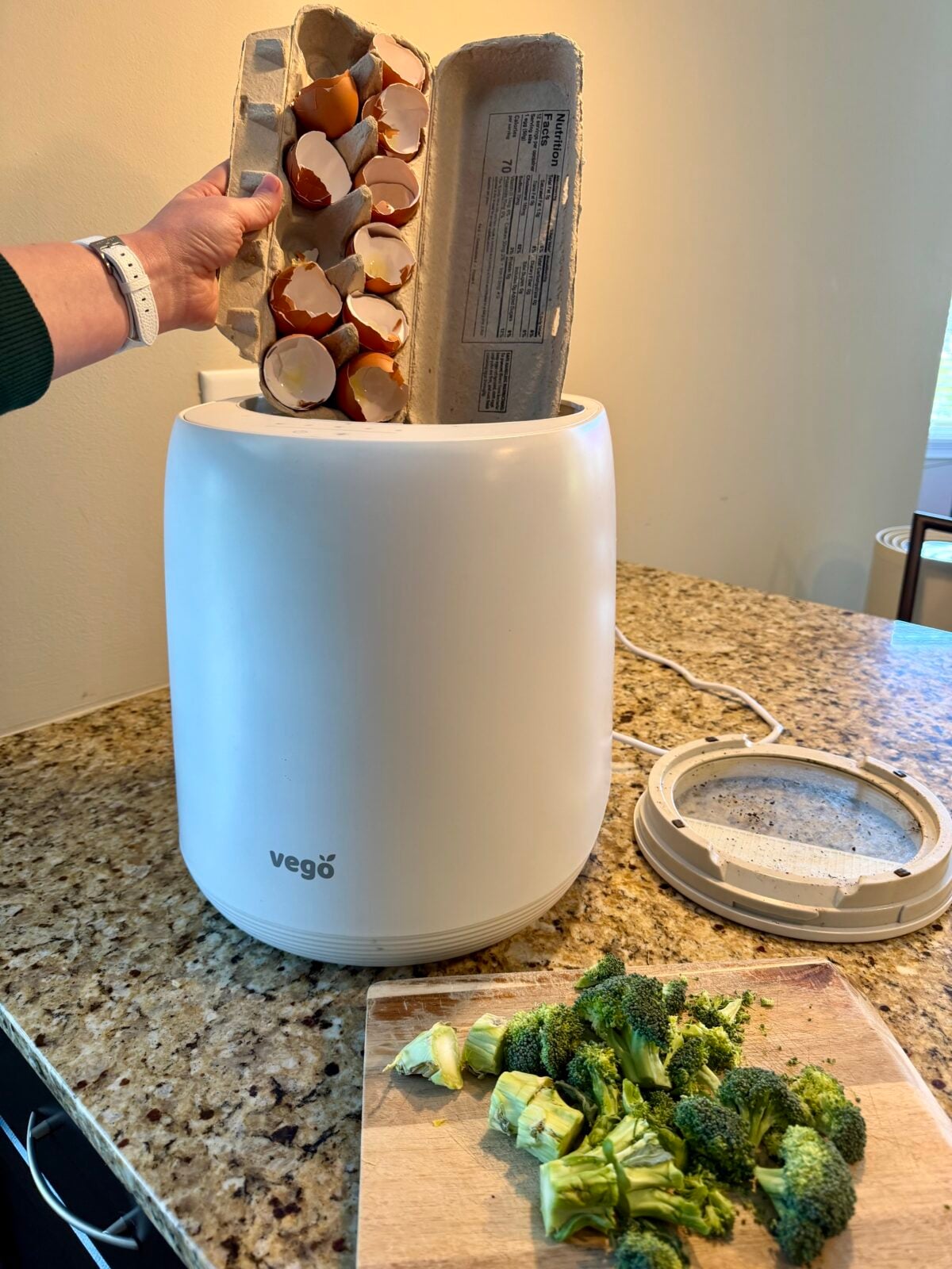 A person inserts an empty egg carton with eggshells into a white Vego compost bin on a kitchen counter. Next to the bin is a cutting board with chopped broccoli. The bin lid rests to the side.