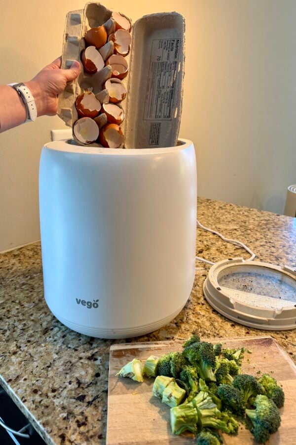 A person inserts an empty egg carton with eggshells into a white Vego compost bin on a kitchen counter. Next to the bin is a cutting board with chopped broccoli. The bin lid rests to the side.