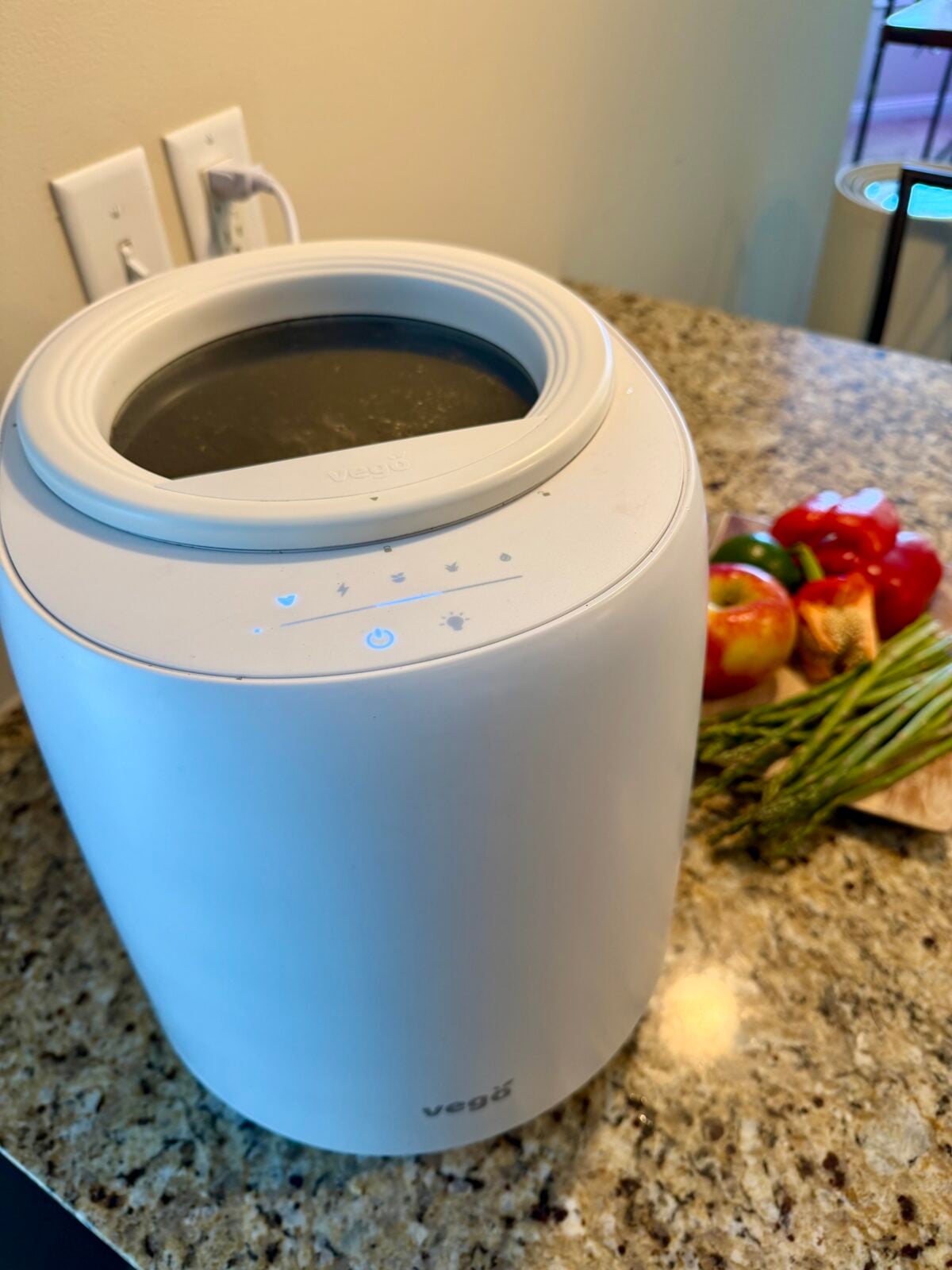 A white kitchen appliance sits on a granite countertop with lights on its control panel. Nearby, there are fresh vegetables including asparagus, bell pepper, tomato, and a few bread pieces.