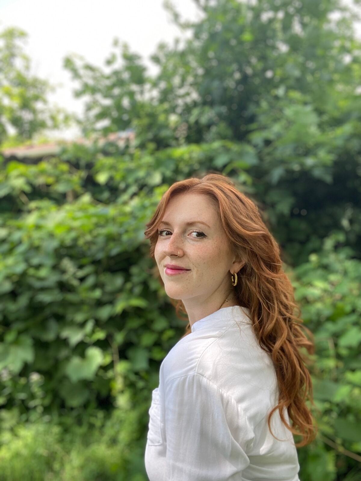 A woman with long red hair and a white shirt stands outdoors in front of dense green foliage, looking over her shoulder at the camera with a slight smile.