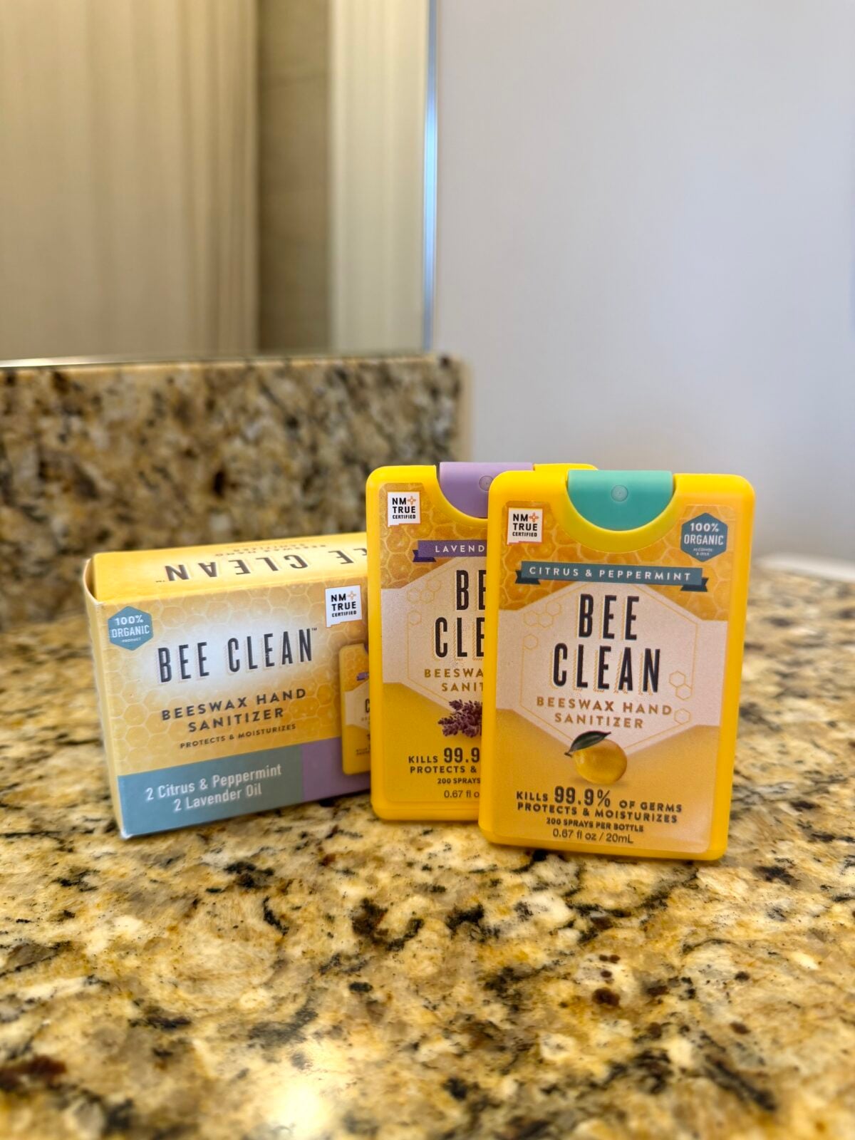 Two yellow containers and one yellow box of Bee Clean Beeswax Hand Sanitizer are placed on a granite bathroom countertop with a mirror and white wall visible in the background.