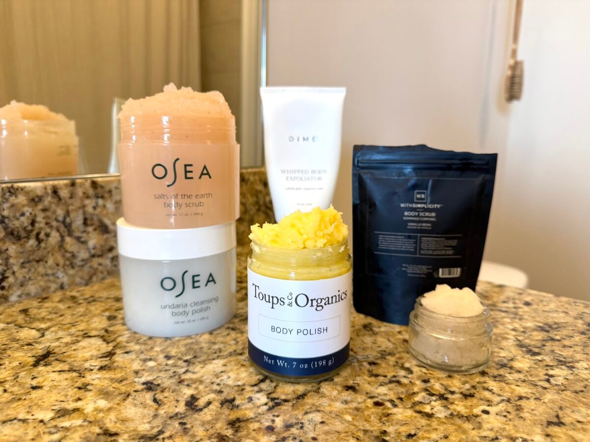 Several body scrubs and polishes in jars and tubes are displayed on a bathroom counter with a granite surface, with a mirror and part of a toothbrush visible in the background.