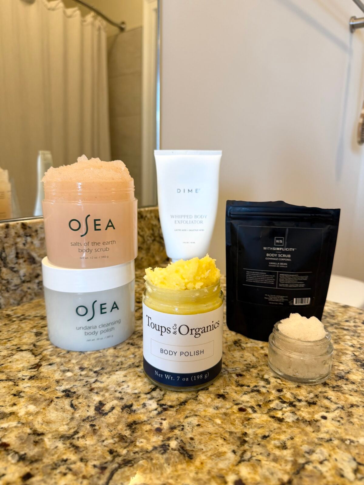 Five body scrub and exfoliation products, including OSEA jars, a DIME tube, a Toups & Co Organics jar, and a black pouch, arranged on a bathroom countertop near a sink and mirror.
