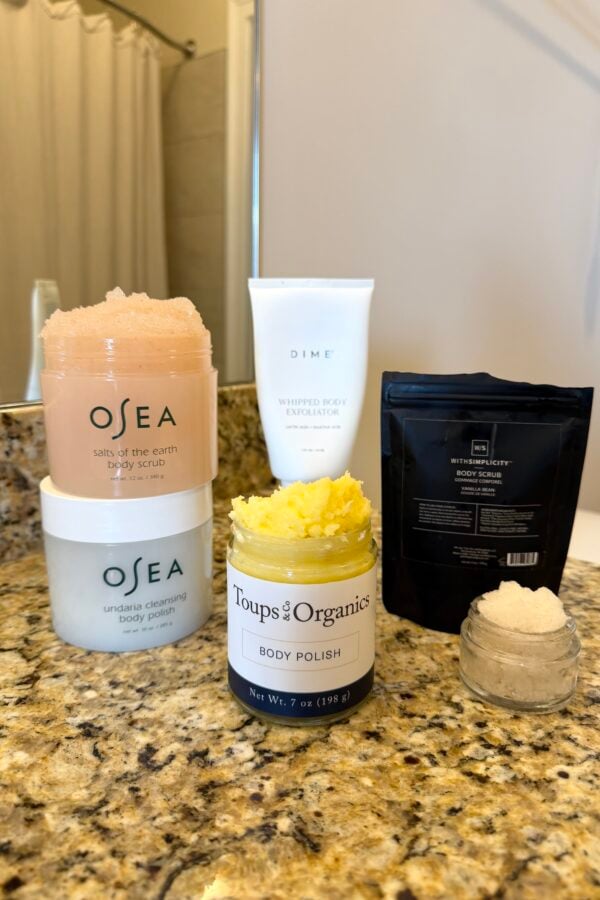 Five body scrub and exfoliation products, including OSEA jars, a DIME tube, a Toups & Co Organics jar, and a black pouch, arranged on a bathroom countertop near a sink and mirror.