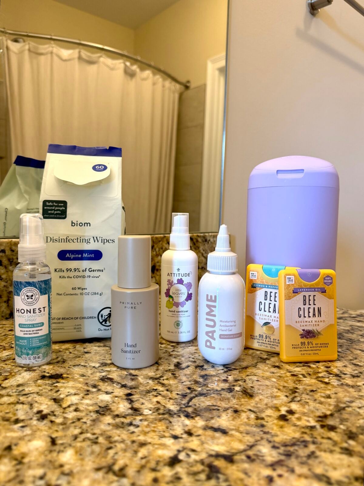 Seven hand sanitizers and disinfecting wipes are arranged on a granite bathroom counter with a purple dispenser. A shower curtain and beige walls are visible in the background.