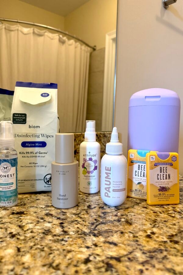 Seven hand sanitizers and disinfecting wipes are arranged on a granite bathroom counter with a purple dispenser. A shower curtain and beige walls are visible in the background.