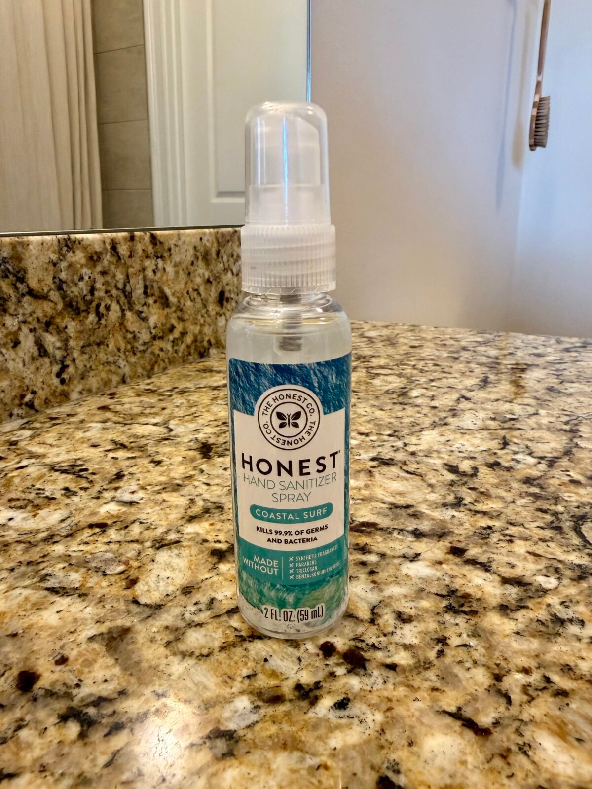 A bottle of Honest Hand Sanitizer Spray, Coastal Surf scent, sits on a granite bathroom countertop. The label indicates it kills 99.9% of germs and is made without harsh chemicals. The bottle contains 2 fl oz.