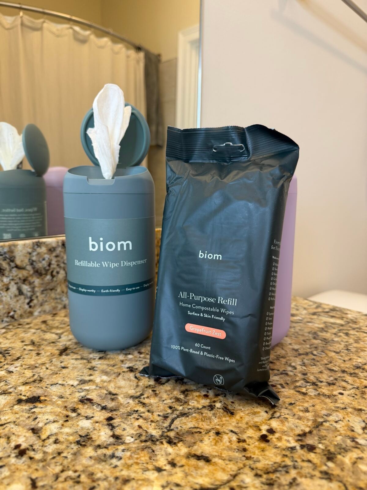 A biom refillable wipe dispenser with a white wipe sticking out sits on a bathroom counter next to a black package labeled biom All-Purpose Refill, containing 60 home compostable wipes.