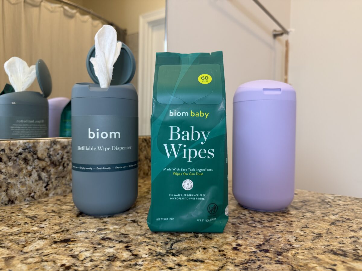 Three containers are on a bathroom counter: a gray biom refillable wipe dispenser with a white wipe, a green bag labeled biom baby Baby Wipes, and a closed pale purple dispenser. A mirror and towel rack are in the background.
