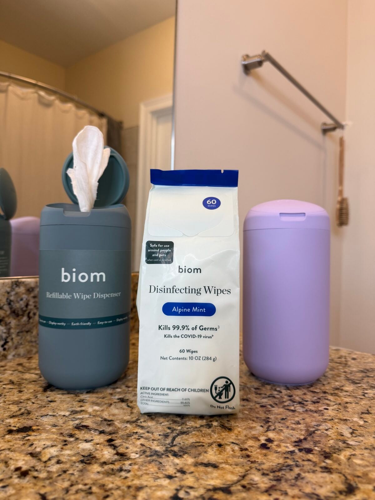 A blue biom refillable wipe dispenser, a pack of biom disinfecting Alpine Mint wipes, and a lavender container sit on a bathroom countertop with a sink, mirror, and shower curtain visible in the background.