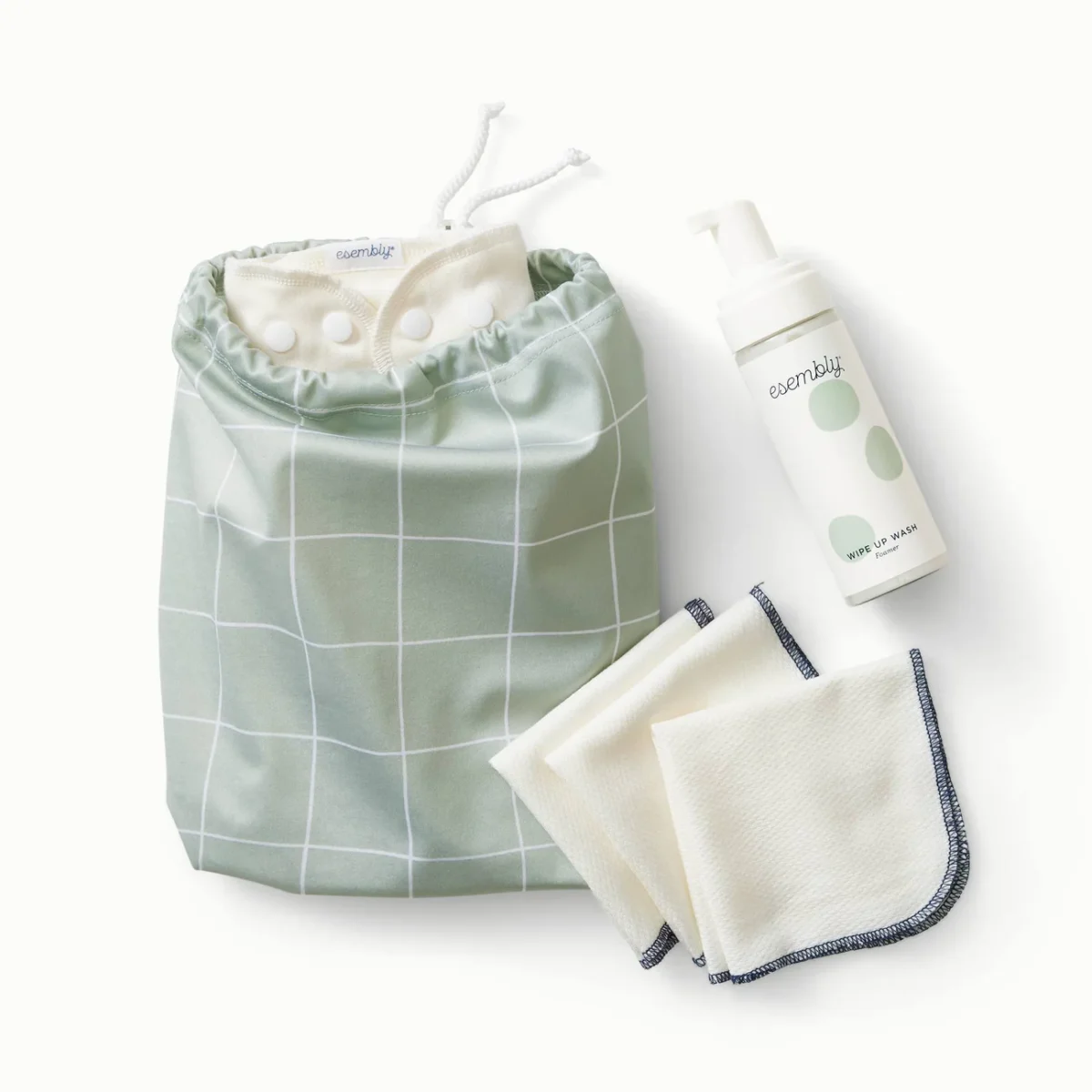 A green and white checkered wet bag, two white cloth wipes with dark edges, a folded white cloth, and a bottle of foaming wash labeled “Esembly” are displayed on a neutral background.