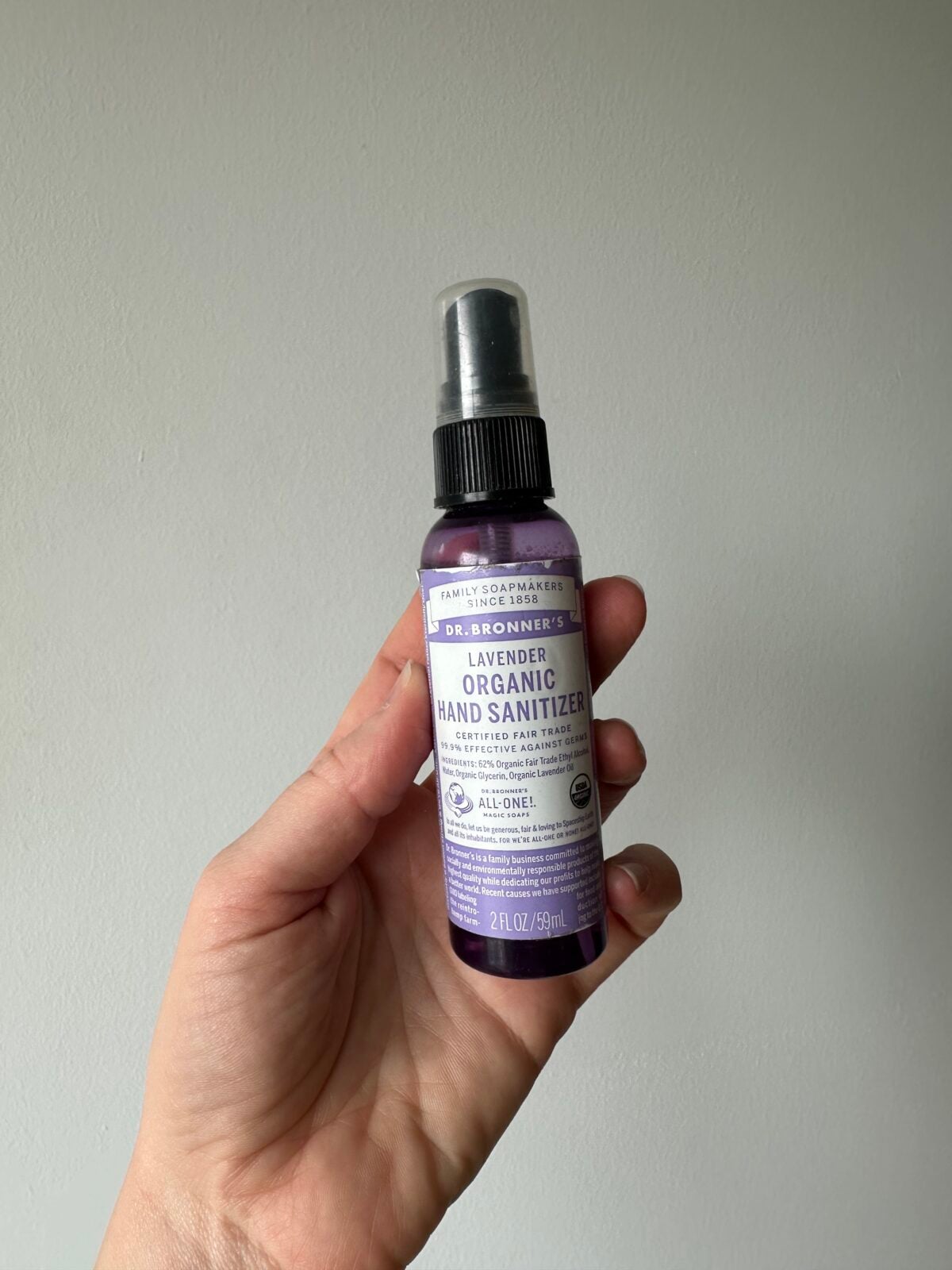 A hand holds a 2 fl oz bottle of Dr. Bronners Lavender Organic Hand Sanitizer against a plain light gray background.