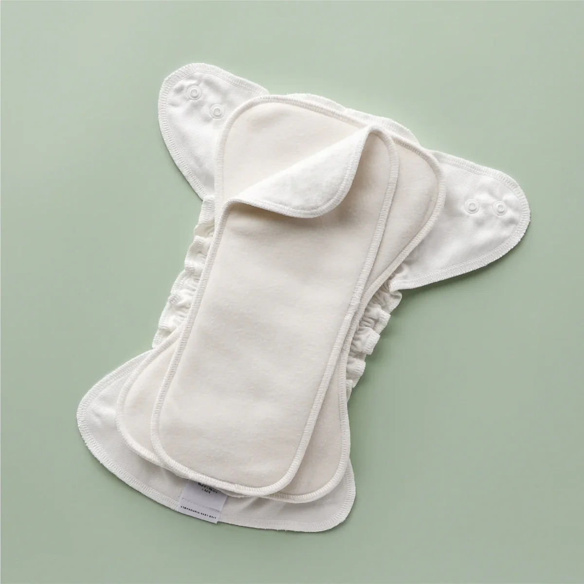 A reusable cloth diaper with snap buttons and absorbent inserts is displayed on a light green background. The diaper is off-white and layered to show different parts, including an extra liner on top.