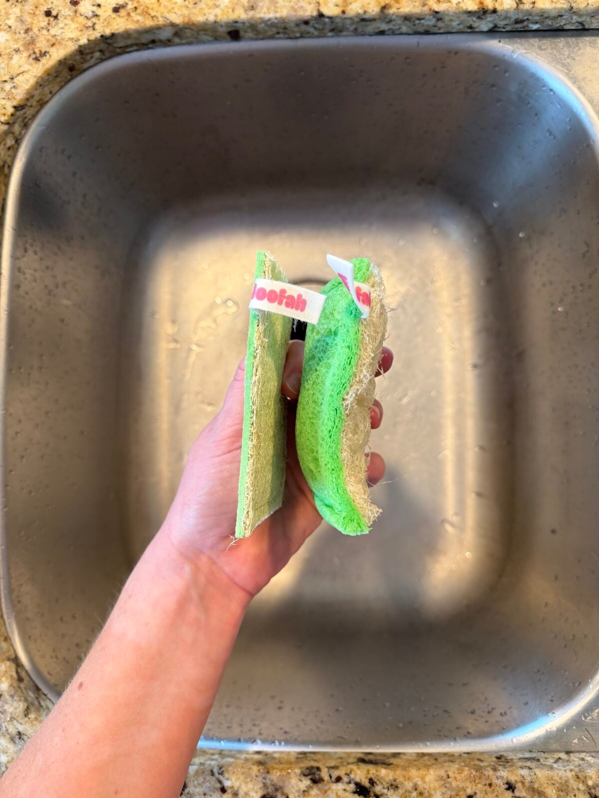 A hand holds two green loofah sponges over a stainless steel sink. One sponge has a small label reading “loofah” and both have a rough-textured surface on one side. The sink and granite countertop are visible.