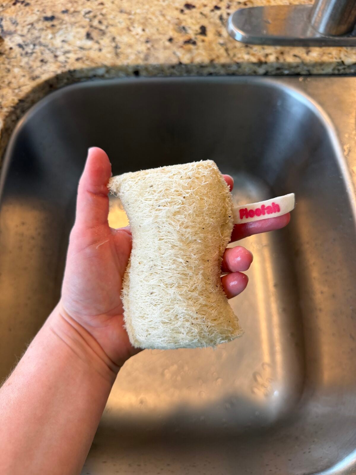 A hand holds a natural loofah sponge with a pink and white tag labeled “Fleelah” above a kitchen sink, with part of a granite countertop visible.