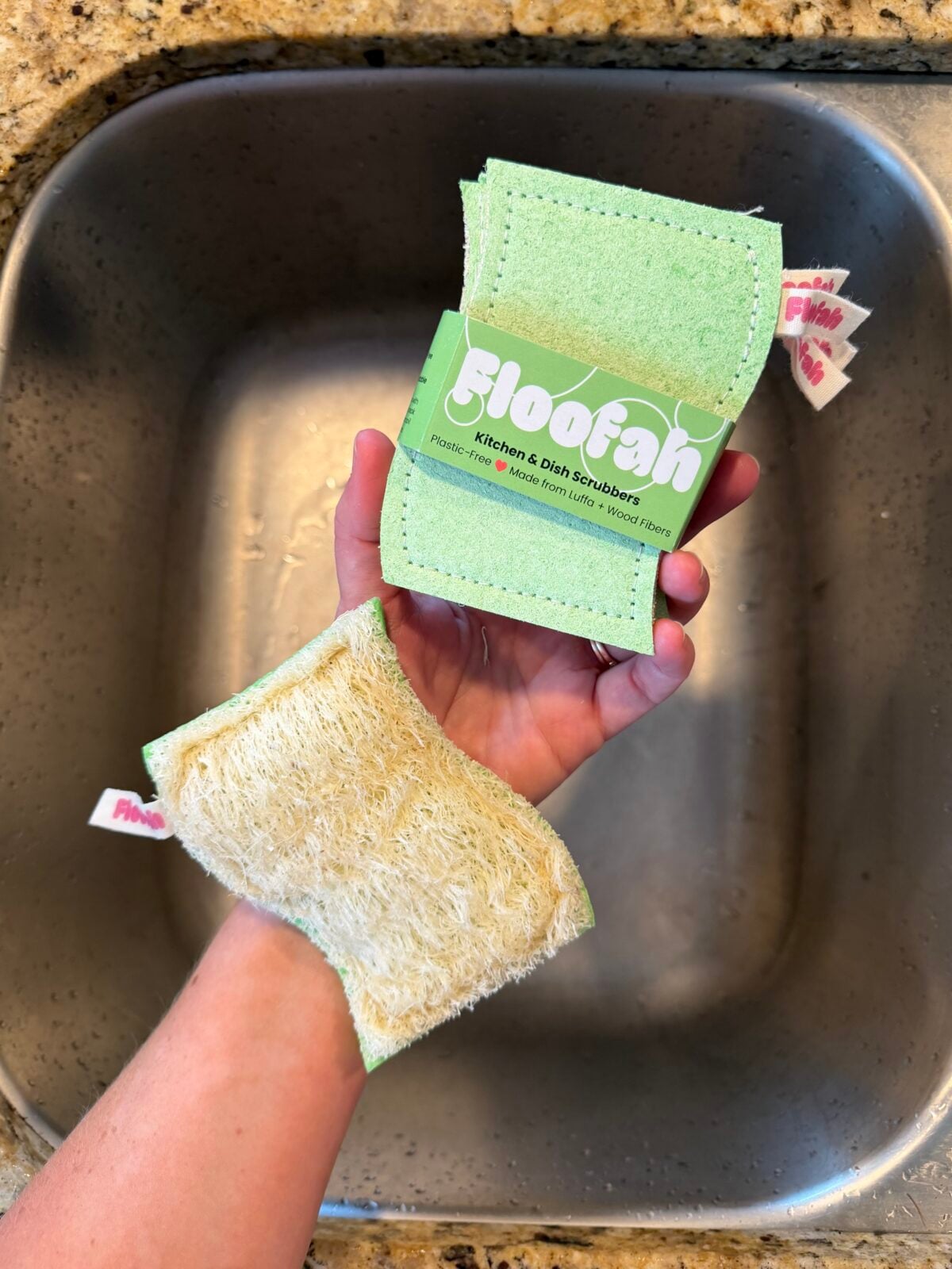 A hand holds two Floofah kitchen cloth scrubbers over a metal sink. One scrubber shows the green label side, while the other displays the textured, natural scrub side. Both have small branded tags attached.