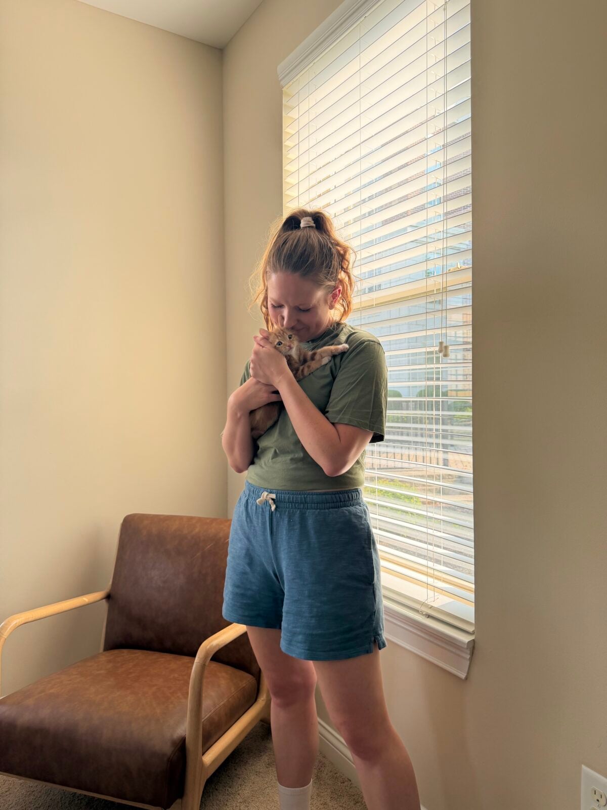 A person stands in a sunlit room by a window, wearing a green shirt and blue shorts, holding a small animal close to their face. A wooden chair with a brown seat is placed nearby.