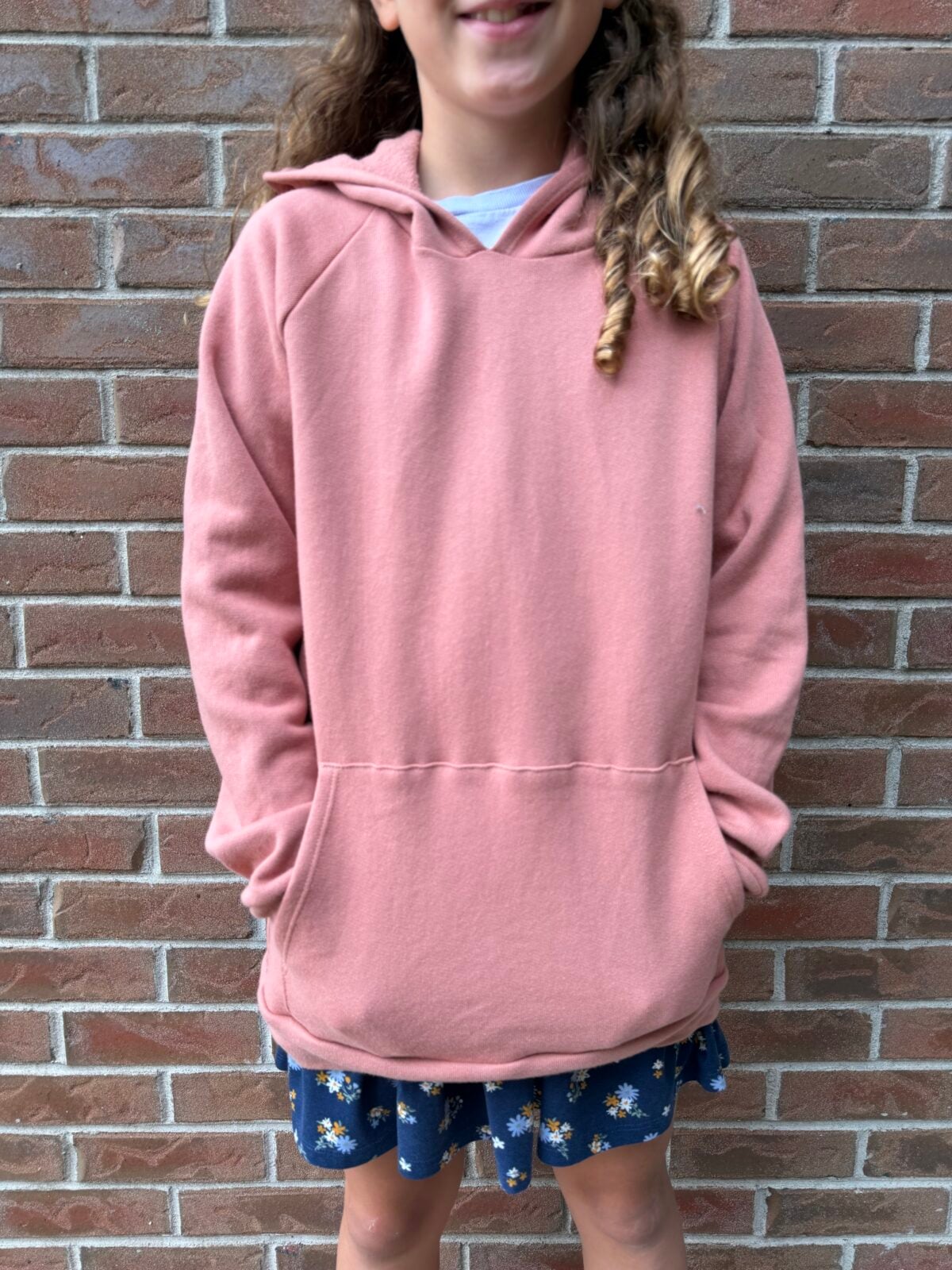A person with long wavy hair stands against a brick wall, wearing a pink hoodie and a blue dress with white flowers. The persons hands are in the pocket of the hoodie and their face is partially out of frame.