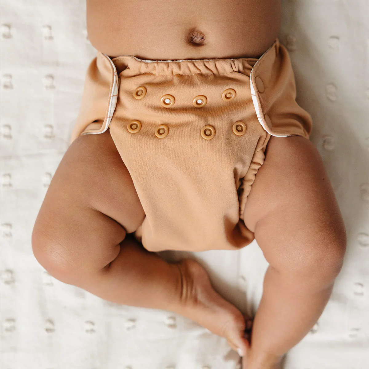 A baby lies on a light-colored blanket, wearing a tan cloth diaper with multiple buttons on the front. Only the babys torso and legs are visible in the image.