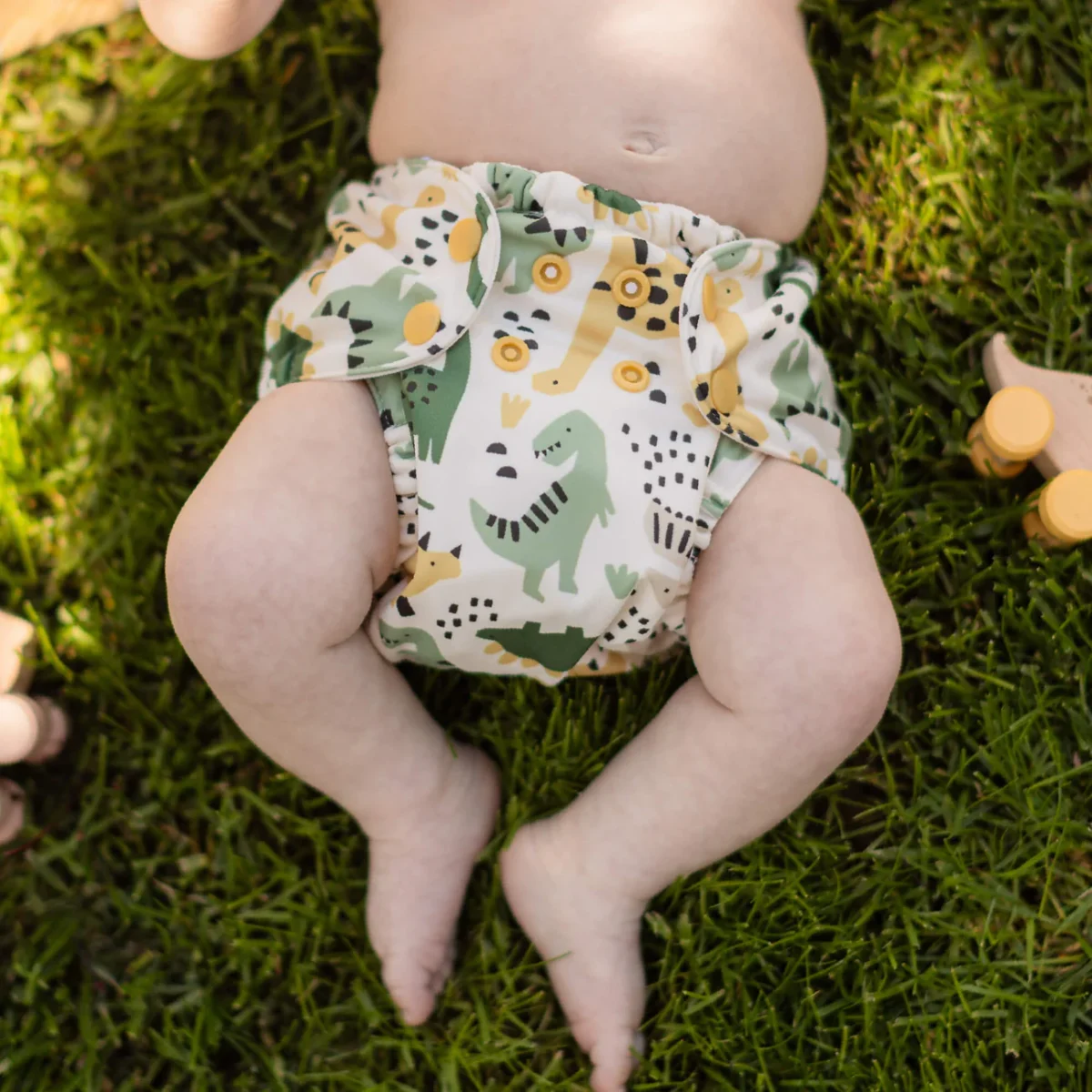 A baby lies on green grass wearing a white cloth diaper with colorful dinosaur and animal prints. Wooden toys are partially visible around the baby. The baby’s torso and legs are shown, but their face is not visible.