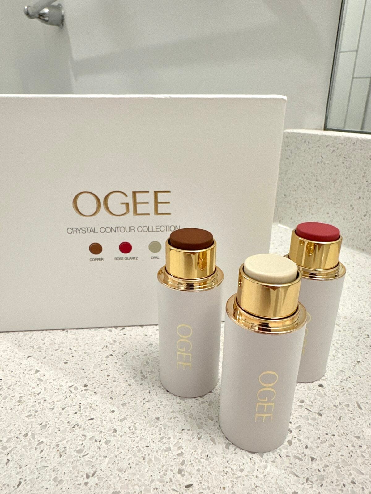 Three OGEE cosmetic sticks with gold accents stand on a countertop in front of a white OGEE Crystal Contour Collection box. The makeup shades are brown, cream, and pink. A sink and mirror are visible in the background.