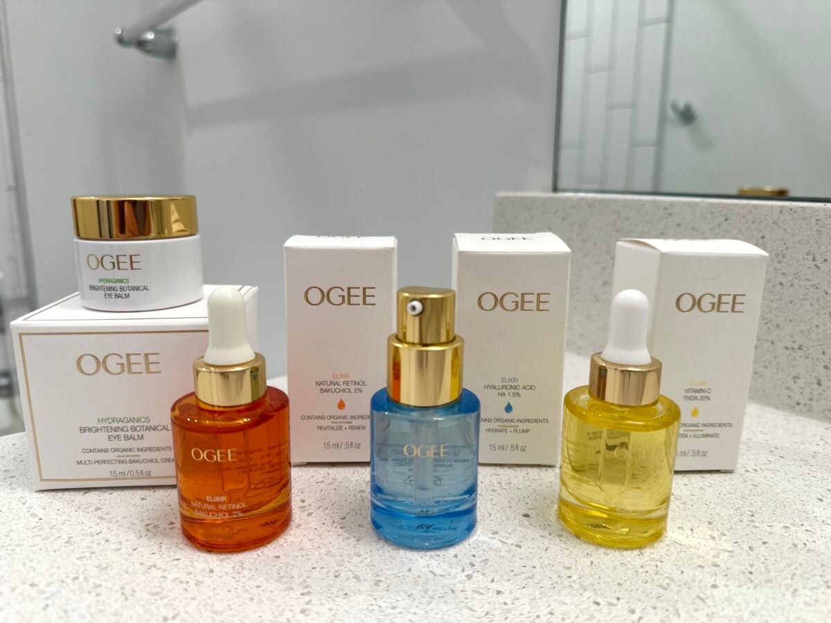 Three OGEE serum bottles—orange, blue, and yellow—stand in front of their matching white boxes on a bathroom counter, next to a small white jar, mirror, and faucet, with a white tiled wall in the background.