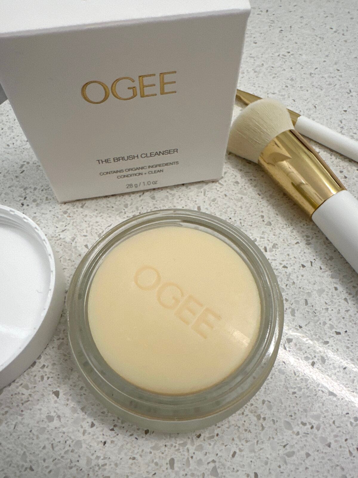 A round container of OGEE brush cleanser with embossed branding sits open on a countertop. The lid is nearby. A white and gold makeup brush and the products box are also visible in the background.