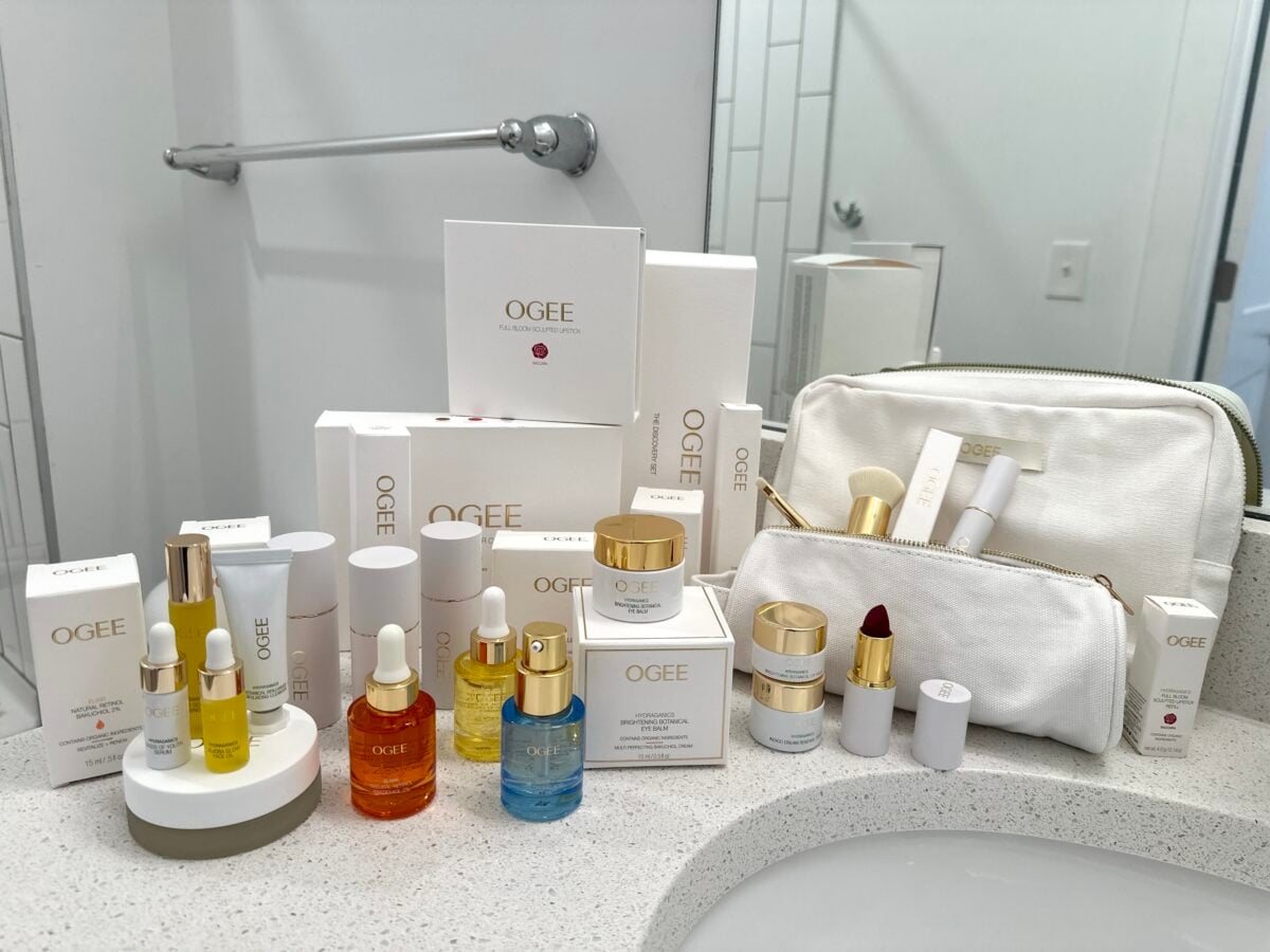 A collection of OGEE skincare and beauty products, including creams, serums, and lip balms, arranged on a bathroom counter with a white zip pouch near a sink and a wall mirror.